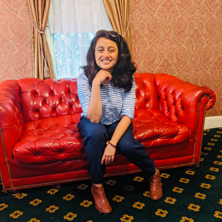 Aditi Patel is the Founder and Designer behind POESHAQ. She is sitting on a red chesterfield couch.