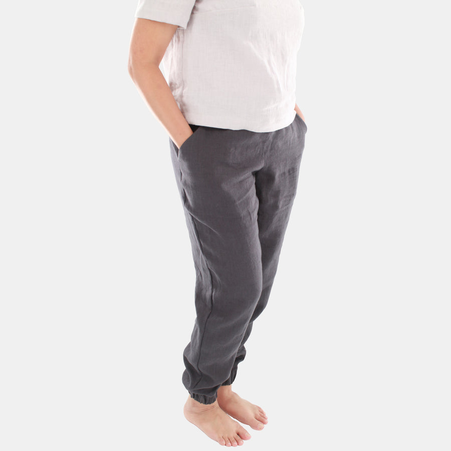 Female model wearing Terra Tapered Pant in Midweight Linen. The pant has an elastic waist and cuffs with front side pockets. Color of the fabric is anthracite grey.