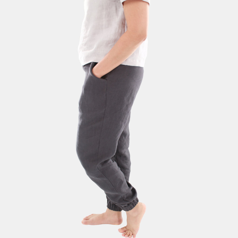 Side view of a female model wearing Terra Tapered Pant in Midweight Linen. The pant has an elastic waist and cuffs with front side pockets. Color of the fabric is anthracite grey.