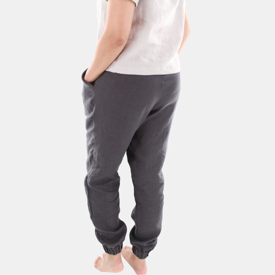 Back view of a female model wearing Terra Tapered Pant in Midweight Linen. The pant has an elastic waist and cuffs with front side pockets. Color of the fabric is anthracite grey.