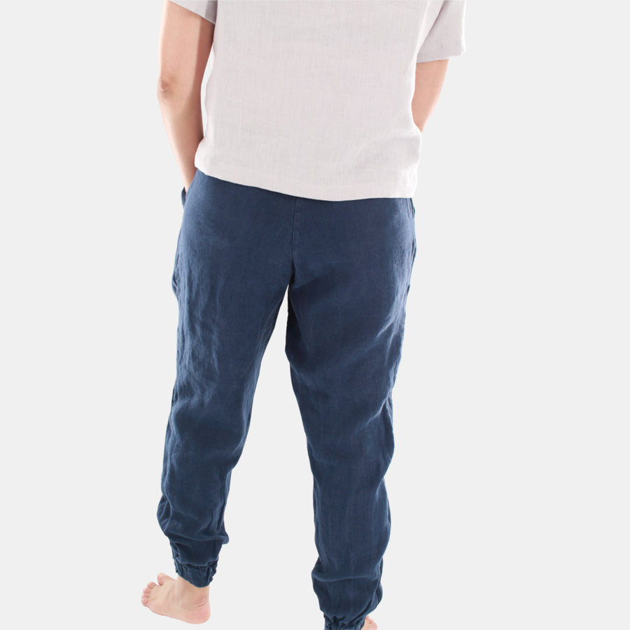 Back view of a female model wearing Terra Tapered Pant in Midweight Linen. The pant has an elastic waist and cuffs with front side pockets. Color of the fabric is navy.