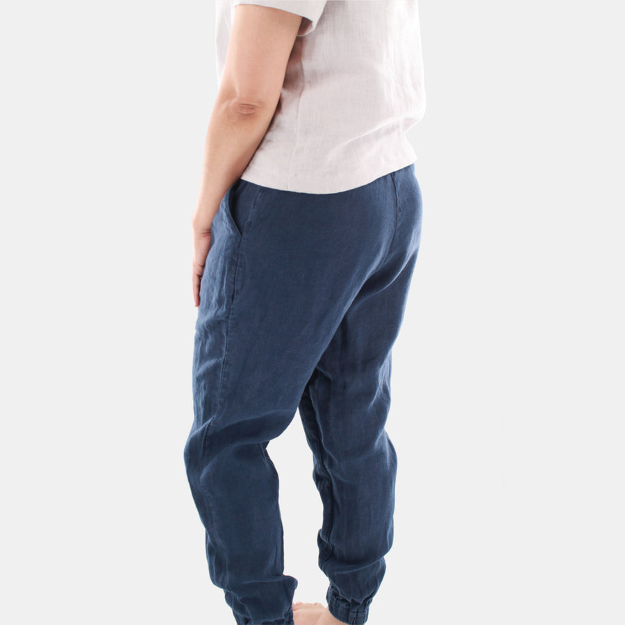 Tilted back view of a female model wearing Terra Tapered Pant in Midweight Linen. The pant has an elastic waist and cuffs with front side pockets. Color of the fabric is navy.