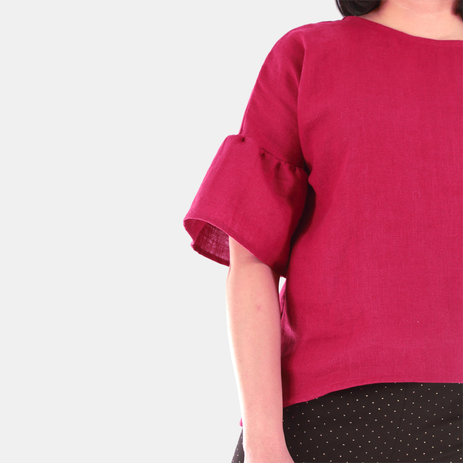 Half close-up of a female model wearing womens willow bell sleeve top in Midweight Linen. Hem finishing is high-low from front to back. Color of the top is Burgundy.
